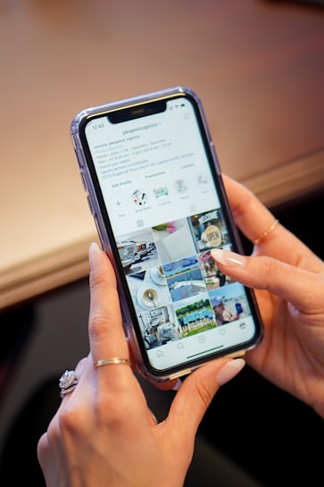 A hand holding a smartphone with an active social media profile page displayed on the screen. The page includes images of people, places, and objects in a grid layout typical of Instagram. The person has manicured nails and is wearing a ring.
