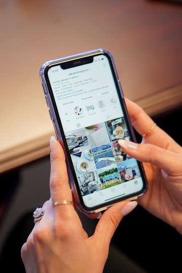 A hand holding a smartphone with an active social media profile page displayed on the screen. The page includes images of people, places, and objects in a grid layout typical of Instagram. The person has manicured nails and is wearing a ring.