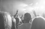 A crowd of people is gathered at a concert, with several individuals raising their hands in a peace sign. The stage is brightly lit with spotlights, creating a festive atmosphere. The image is in black and white, adding a timeless feel.