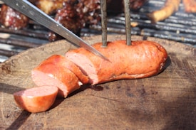 A cooked sausage is cut into slices using a knife on a wooden cutting board. Metal skewers hold the sausage in place. The background shows a grill with other food items.