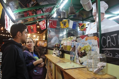 Two people stand at a food stall, engaging with a vendor wearing a chef's uniform. The stall features various condiments, jars, and bottles, with decorative flags hanging above. A menu sign displays food options.