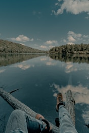 person in black and white sneakers sitting on wooden dock over the river during daytime