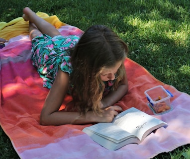 girl in pink dress reading book on green grass during daytime