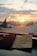 A warm, soft-focus sunset is seen through an airport window, with the silhouettes of airplanes just visible. In the foreground, a boarding pass and a blue passport lay on the windowsill, adding to the theme of travel and anticipation.