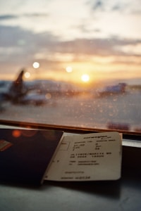 A warm, soft-focus sunset is seen through an airport window, with the silhouettes of airplanes just visible. In the foreground, a boarding pass and a blue passport lay on the windowsill, adding to the theme of travel and anticipation.