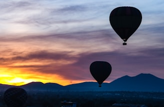 Hot air balloons float in the sky during sunrise. The horizon shows an array of mountains and a sky painted with warm tones of orange and yellow blending into darker shades of blue and black. The silhouette of the balloons contrasts against the brightening sky.