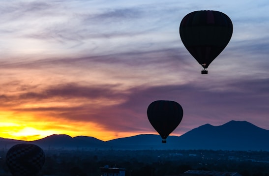 Hot air balloons float in the sky during sunrise. The horizon shows an array of mountains and a sky painted with warm tones of orange and yellow blending into darker shades of blue and black. The silhouette of the balloons contrasts against the brightening sky.