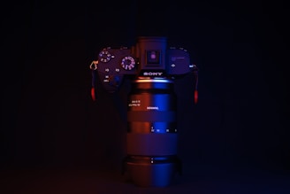 A camera with a lens stands against a dark background, illuminated with red and blue lighting. The brand names SONY and TAMRON are visible on the camera body and lens, respectively. The lighting highlights the contours and buttons of the camera.