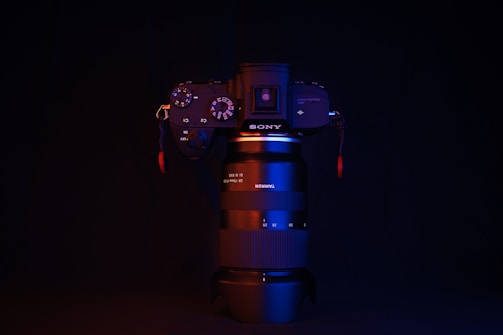 A camera with a lens stands against a dark background, illuminated with red and blue lighting. The brand names SONY and TAMRON are visible on the camera body and lens, respectively. The lighting highlights the contours and buttons of the camera.