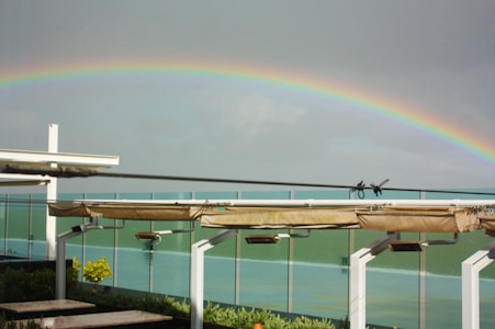 A vibrant rainbow arches across a cloudy sky above a modern structure with white beams and glass panels. Below the rainbow, a garden area with lush greenery and a few yellow flowers is visible.