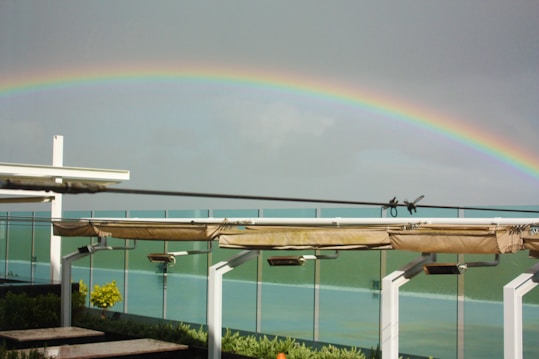 A vibrant rainbow arches across a cloudy sky above a modern structure with white beams and glass panels. Below the rainbow, a garden area with lush greenery and a few yellow flowers is visible.