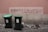 A pink wall with graffiti written in Portuguese, reading 'Acorda! Deus é um sonho! Não acredito.' Below the graffiti are two green garbage bins placed on a stone pavement. Some debris and rocks lie on the ground near the wall.