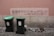A pink wall with graffiti written in Portuguese, reading 'Acorda! Deus é um sonho! Não acredito.' Below the graffiti are two green garbage bins placed on a stone pavement. Some debris and rocks lie on the ground near the wall.