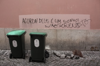 A pink wall with graffiti written in Portuguese, reading 'Acorda! Deus é um sonho! Não acredito.' Below the graffiti are two green garbage bins placed on a stone pavement. Some debris and rocks lie on the ground near the wall.