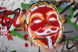 Vibrant street art featuring a stylized mask painted in red, yellow, and white, with a background of abstract graffiti. The mask has an enigmatic smile and is set against a wall with overgrown plants at the bottom.