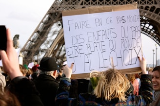 A crowd is gathered near a large structure resembling the Eiffel Tower. A person holds up a sign with French text, and several other people are visible holding signs in the background.