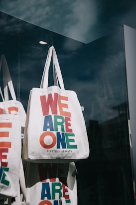 Multiple white tote bags with bold, colorful text in red, blue, and green are displayed in a shop window. The phrase 'WE ARE ONE UNITED. ALWAYS' is printed on the bags. The setting includes reflections on the glass, suggesting an outdoor environment.