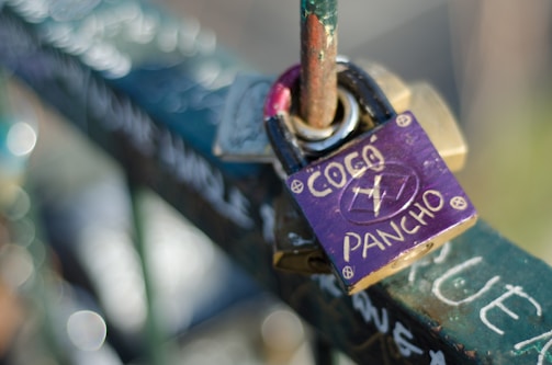 A purple padlock with the words 'COCO' and 'PANCHO' along with some symbols on it is attached to a metal railing. The railing and lock are decorated with graffiti and the background is out of focus, showing light bokeh.