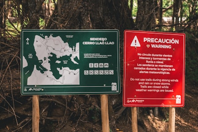 Two signs are posted on wooden stakes amidst a forest background. The left sign is green and displays a map of the Cerro Llao Llao hiking trail, complete with path outlines, waypoints, and details such as difficulty and estimated walking time. The right sign, red in color, provides a warning in both Spanish and English about avoiding the trails during adverse weather conditions like strong winds and storms.