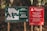 Two signs are posted on wooden stakes amidst a forest background. The left sign is green and displays a map of the Cerro Llao Llao hiking trail, complete with path outlines, waypoints, and details such as difficulty and estimated walking time. The right sign, red in color, provides a warning in both Spanish and English about avoiding the trails during adverse weather conditions like strong winds and storms.