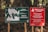Two signs are posted on wooden stakes amidst a forest background. The left sign is green and displays a map of the Cerro Llao Llao hiking trail, complete with path outlines, waypoints, and details such as difficulty and estimated walking time. The right sign, red in color, provides a warning in both Spanish and English about avoiding the trails during adverse weather conditions like strong winds and storms.