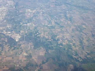 An aerial view of a rural landscape with a patchwork of agricultural fields. The area is interspersed with patches of forests and winding rivers. There are visible roads and small clusters of buildings, indicating towns or settlements.