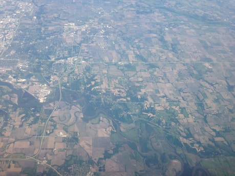 An aerial view of a rural landscape with a patchwork of agricultural fields. The area is interspersed with patches of forests and winding rivers. There are visible roads and small clusters of buildings, indicating towns or settlements.