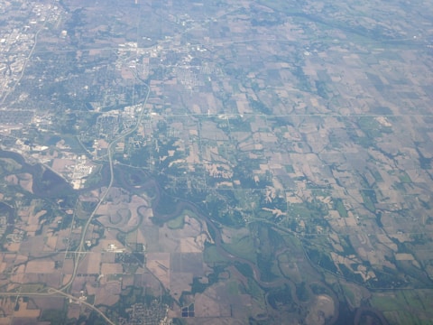 An aerial view of a rural landscape with a patchwork of agricultural fields. The area is interspersed with patches of forests and winding rivers. There are visible roads and small clusters of buildings, indicating towns or settlements.