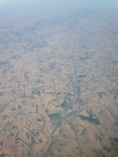 An aerial view of expansive farmland crisscrossed by roads and patches of greenery. The landscape is covered with fields of varying sizes and shapes, displaying shades of brown and green. A river meanders through the terrain, providing a natural contrast to the man-made agricultural patterns.