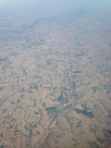 An aerial view of expansive farmland crisscrossed by roads and patches of greenery. The landscape is covered with fields of varying sizes and shapes, displaying shades of brown and green. A river meanders through the terrain, providing a natural contrast to the man-made agricultural patterns.