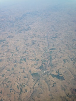 An aerial view of expansive farmland crisscrossed by roads and patches of greenery. The landscape is covered with fields of varying sizes and shapes, displaying shades of brown and green. A river meanders through the terrain, providing a natural contrast to the man-made agricultural patterns.