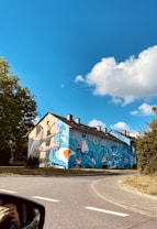 A building with a vibrant and large mural painted on its side, depicting a person spray painting with dynamic blue, orange, and white abstract designs. The scene is set on a clear day with a bright blue sky and fluffy clouds, surrounded by green trees and a quiet street.