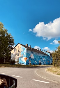 A building with a vibrant and large mural painted on its side, depicting a person spray painting with dynamic blue, orange, and white abstract designs. The scene is set on a clear day with a bright blue sky and fluffy clouds, surrounded by green trees and a quiet street.