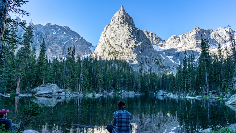 person in red and blue plaid shirt sitting on rock facing lake and snow covered mountain