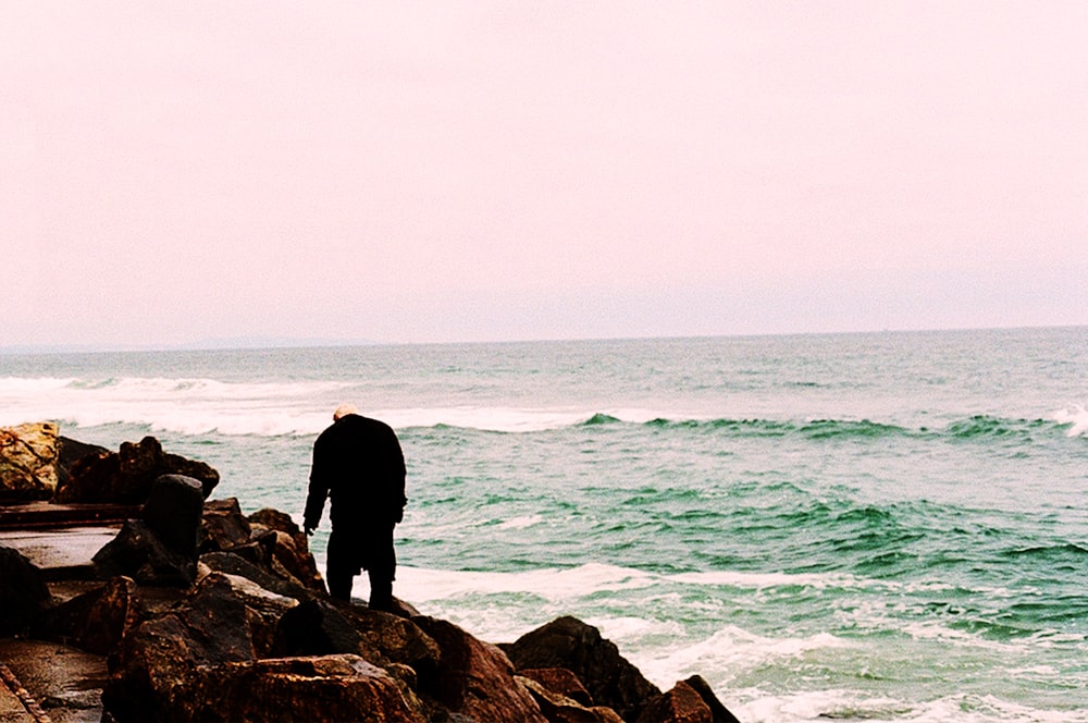 man in black jacket standing on brown rock near sea during daytime