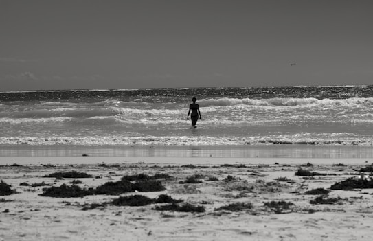 A lone person walks towards the ocean, set against the backdrop of a calm seascape with gentle waves. The beach foreground is scattered with seaweed and the scene is captured in monochrome, giving it a timeless and serene atmosphere.