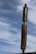 A tall, wooden totem pole with a stylized human face stands prominently against a backdrop of a lightly clouded sky. The totem is weathered and has a rustic appearance, with carved facial features including a prominent nose and mouth. The sky is filled with soft, wispy clouds, creating a serene and expansive atmosphere.