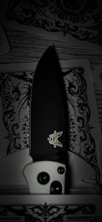 A detailed folding knife with a black blade and a logo visible near the handle rests on intricately designed playing cards, featuring decorative black and white patterns.