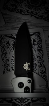 A detailed folding knife with a black blade and a logo visible near the handle rests on intricately designed playing cards, featuring decorative black and white patterns.