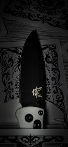 A detailed folding knife with a black blade and a logo visible near the handle rests on intricately designed playing cards, featuring decorative black and white patterns.