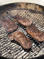 Four pieces of seasoned meat are being grilled on a barbecue. The grill is circular, containing hot coals underneath, and the meat appears to be in the process of cooking with some char marks visible on the surface.