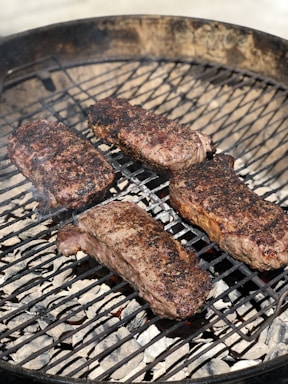 Four pieces of seasoned meat are being grilled on a barbecue. The grill is circular, containing hot coals underneath, and the meat appears to be in the process of cooking with some char marks visible on the surface.