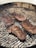 Four pieces of seasoned meat are being grilled on a barbecue. The grill is circular, containing hot coals underneath, and the meat appears to be in the process of cooking with some char marks visible on the surface.