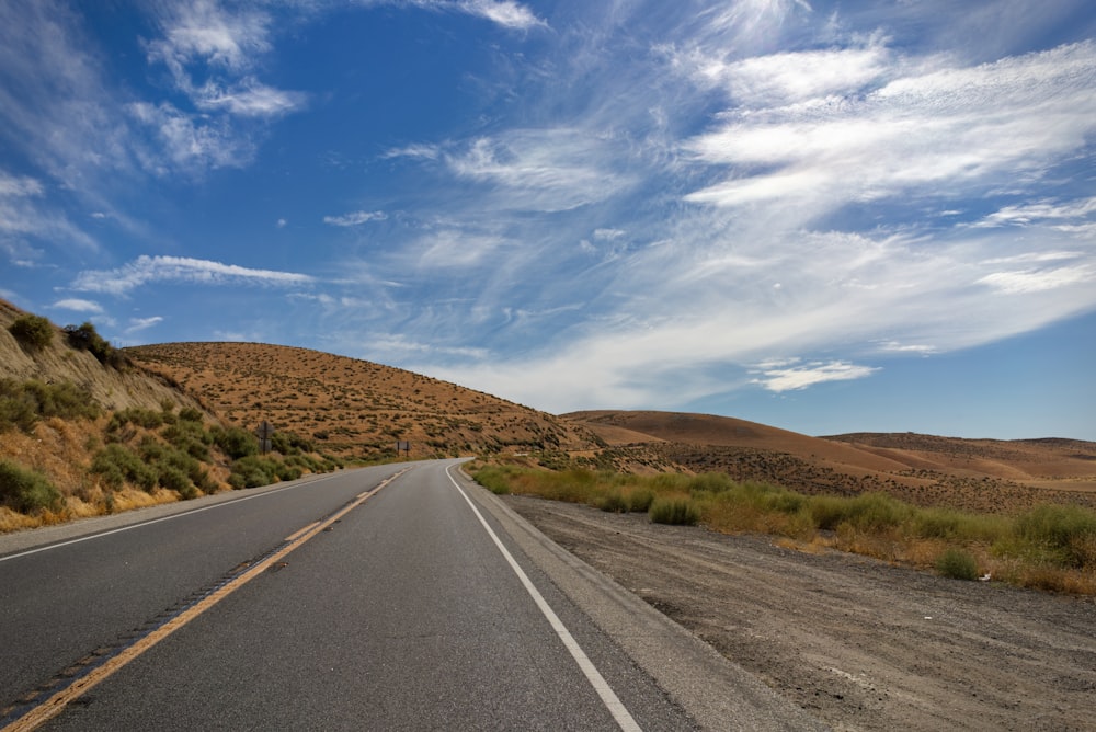 strada asfaltata grigia tra il campo di erba marrone sotto il cielo nuvoloso soleggiato blu e bianco durante il giorno