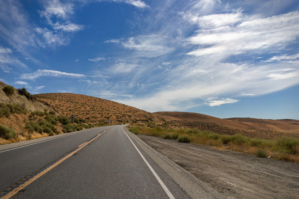 The image depicts a two-lane highway winding through a desert landscape, with a clear blue sky overhead and fluffy white clouds. The road is marked by a yellow center line on its left side, guiding vehicles along their journey. On either side of the road, there are hills covered in dry grass that stretch out into the distance, creating a sense of depth and vastness to the scene.

The perspective of the image is from within a car traveling down the highway, looking towards the horizon where the sky meets the earth. The colors in the photo are predominantly blue, green, and brown, reflecting the natural elements present in the desert landscape.
