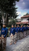 A group of men is standing in a line, wearing traditional blue shirts and patterned sarongs. They are barefoot and appear to be participating in a cultural or ceremonial event. The setting includes trees and buildings with red roofs in the background, under a partly cloudy sky.