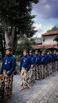 A group of men is standing in a line, wearing traditional blue shirts and patterned sarongs. They are barefoot and appear to be participating in a cultural or ceremonial event. The setting includes trees and buildings with red roofs in the background, under a partly cloudy sky.