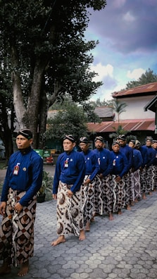 A group of men is standing in a line, wearing traditional blue shirts and patterned sarongs. They are barefoot and appear to be participating in a cultural or ceremonial event. The setting includes trees and buildings with red roofs in the background, under a partly cloudy sky.