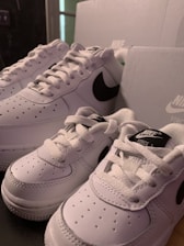 A pair of white Nike sneakers with black swoosh logos are displayed next to matching white shoe boxes. The sneakers appear to be of different sizes, possibly indicating adult and child sizes.