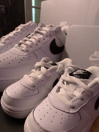 A pair of white Nike sneakers with black swoosh logos are displayed next to matching white shoe boxes. The sneakers appear to be of different sizes, possibly indicating adult and child sizes.